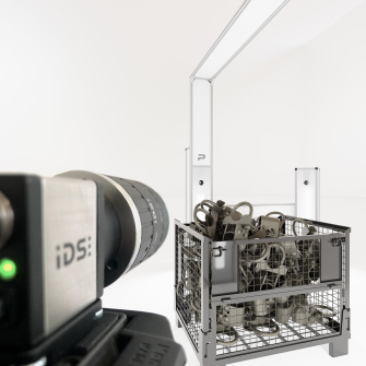 IDS uEye CP Industriekamera, die auf eine Gitterbox mit Metallteilen gerichtet ist, während im Hintergrund eine moderne Beleuchtungs- und Scaneinheit zu sehen ist