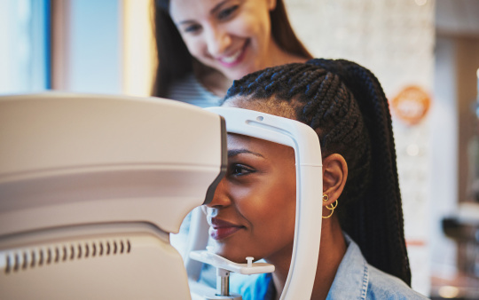 Frau blickt durch Ophthalmologie-Gerät, Optikerin steht dahinter.