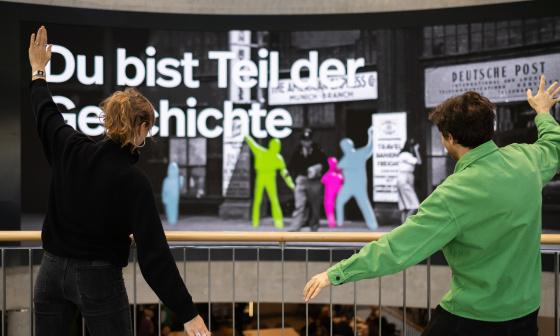 Zwei Personen posieren vor einer großen interaktiven Displaywand mit farbigen Silhouetten