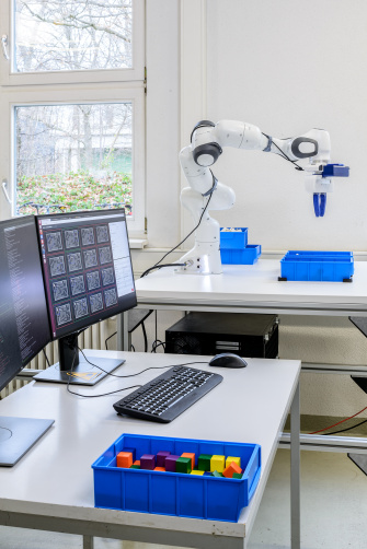 Vor Roboter, der Schrauben aus blauen Behältnis sortiert, befinden sich 2 Bildschirme.