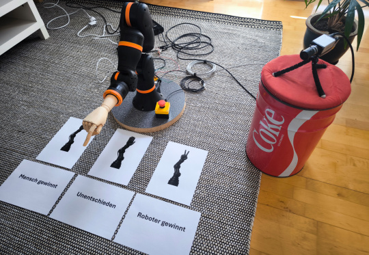 Roboterarm zeigt auf vor ihm liegende Papierblätter, IDS NXT Kamera steht daneben