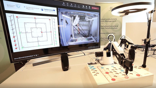 Roboter spielt auf dem Spielbrett Mühle und wird von Lampe beleuchtet, nebendran Bildschirm
