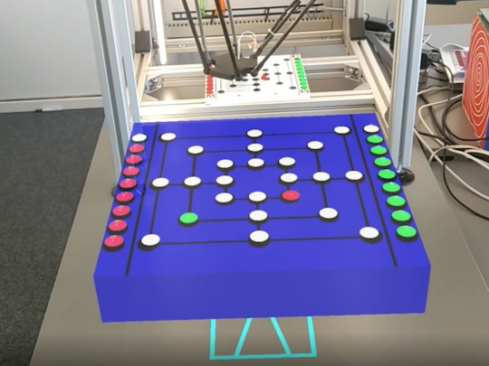 Roboter spielt in Kasten Mühle, vor Kasten das Äquivalent-Spielbrett um gegen den Roboter zu spielen